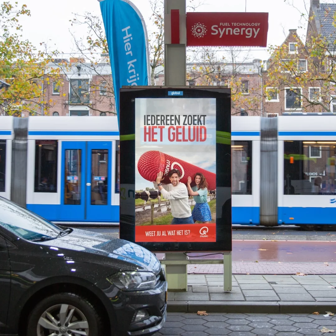 Digitale DOOH-reclame op een Amsterdamse tramhalte met een Qmusic campagne 'IEDEREEN ZOEKT HET GELUID', blauwe tram passeert op de achtergrond, op een herfstachtige dag
